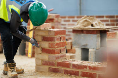 An apprentice in a green hard hat pointing a brick wall