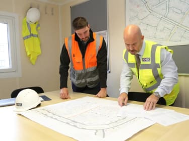 two men in an office wearing safety clothes and looking at a map