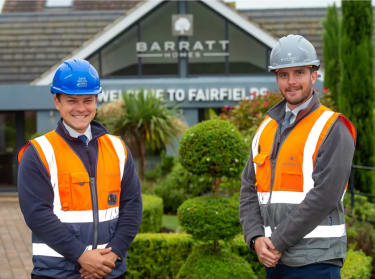 two men in orange high vis vests and hard hats in front of a Barratt Homes sign