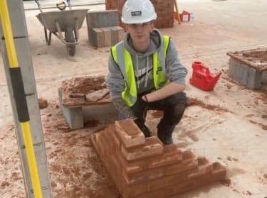 A young man wearing a hi-vis vest squatting behind a half finished brick wall at a construction site