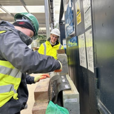 a man wearing safety clothes, laying bricks at a construction site while another man is watching him