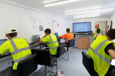 people in hi-vis jackets and hard hards sat at desks learning from laptops