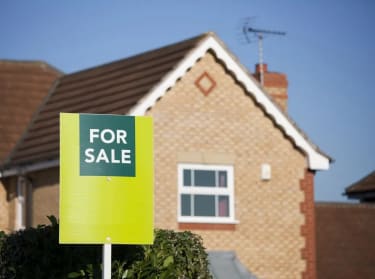 a red-brick house with a green for sale sign in front of it