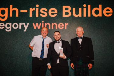 Lyam Gunner smiling at the camera with his Pride in the Job 2024 award alongside CEO Steve Woods and host Al Murray