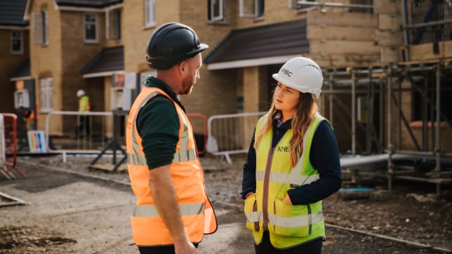 a man and a woman in hi-vis jackets on site in front of some houses talking