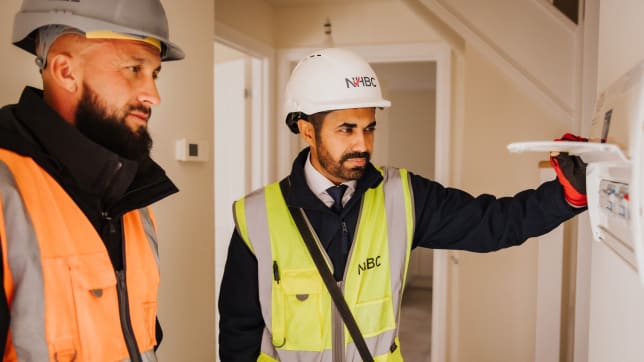 Two men in hi-vis and hard hats inspecting a fusebox inside a house