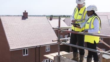 two people in site safety clothing and hard hats stand on scaffolding and look at plans