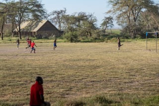 Men playing soccer on a dry grass field with trees and a rustic building in the background.