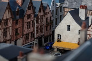 Aerial view of a miniature model depicting European-style buildings, with a focus on a white building with a yellow awning labeled "L'épée Royale" and miniature cars parked nearby.