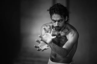 Black and white image of a shirtless man with intense gaze, reaching out with his hands, slightly out of focus against a blurred background.
