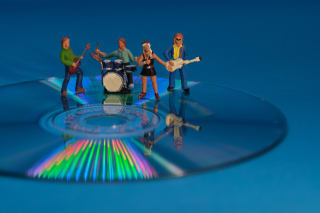 Miniature figurines of a band, including a guitarist, drummer, singer, and bassist, positioned on a CD with a reflective rainbow pattern on a blue background.