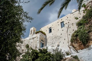 A historic building with whitewashed walls and blue windows is partially obscured by greenery, set against a dramatic sky. The structure features a distinctive bell tower and terraced plants.