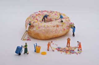 A large donut topped with white icing and colorful sprinkles, surrounded by miniature figurines extracting and collecting the sprinckles.