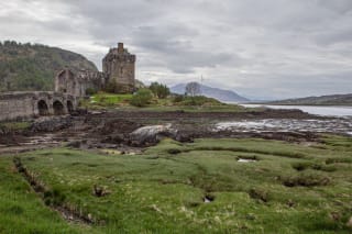 An old stone castle with an arched bridge overlooking a coastal landscape with grassy shores and distant mountains under a cloudy sky.