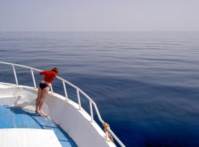 Woman wearing a red jacket, standing at the bow of a boat, looking out over the calm blue sea.