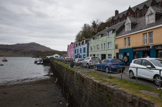 Colorful buildings along a seaside street with parked cars, a stone wall, and boats on the water with a mountainous backdrop.