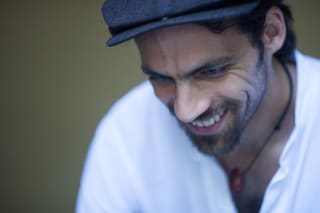 A smiling man wearing a flat cap and a white shirt, looking downwards. The background is out of focus.