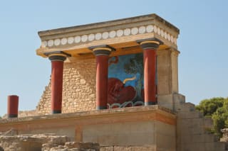 A partial view of a Minoan temple reconstruction featuring red columns, a colorful fresco of a bull, and stone walls under a clear blue sky.