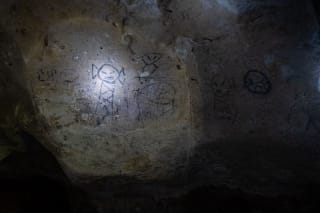 The image shows ancient rock art depicting various figures, illuminated by a flashlight. The carvings include humanoid shapes and other abstract designs on a textured stone surface.