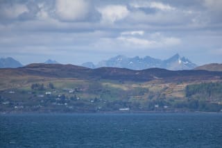 A coastal landscape with a body of water in the foreground, a village amidst rolling hills in the middle, and a mountain range with snowy peaks in the background, under a cloudy sky.