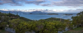 A panoramic view featuring a coastline with several small islands in the ocean, surrounded by lush greenery and distant mountains under a cloudy sky.
