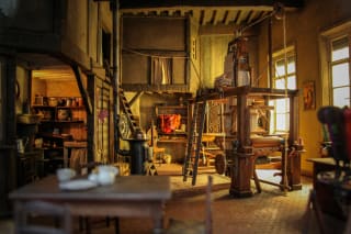 A detailed miniature of an old-fashioned workshop interior with a spinning wheel, shelves with pottery, a fireplace, and a ladder leading to an upper level.