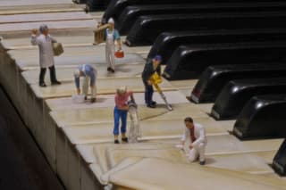 A miniature scene on a piano keyboard depicts small figures engaged in various cleaning and maintenance tasks, such as painting, mopping, and using tools.