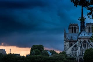 Notre-Dame Cathedral in Paris against a backdrop of a stormy sky with a glimpse of the Eiffel Tower in the distance.