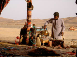 An Egyptian man in traditional dress preparing tea in a tent, against a desert backdrop, with a camel in the background.