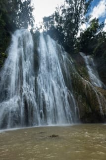 A majestic waterfall cascading down rocky cliffs into a muddy pool, surrounded by lush greenery.