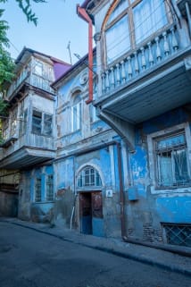 A narrow street with weathered blue buildings, including one with a wooden balcony and decorative architecture.