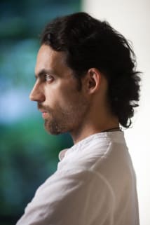 Side profile of a man with dark hair wearing a white shirt, against a blurred background.