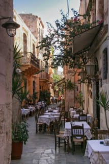 Narrow cobblestone street lined with outdoor café tables and chairs, surrounded by historic buildings with balconies and plants in pots.