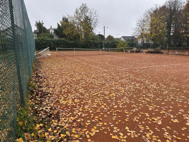 Un terrain de tennis qui semble à l'abandon, couvert de feuilles et avec la chaise d'arbitrage renversée sur une chaise de jardin