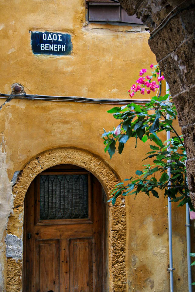 An old, rustic wooden door set within a textured, yellow ochre wall. Above the door is a street sign with Greek text. Pink flowers and green leaves partially obscure the view.