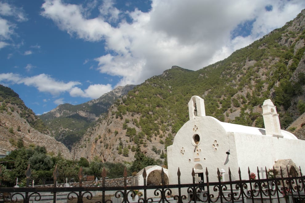 A white church with a bell tower surrounded by a mountainous landscape under a partly cloudy sky. A decorative fence is visible in the foreground.