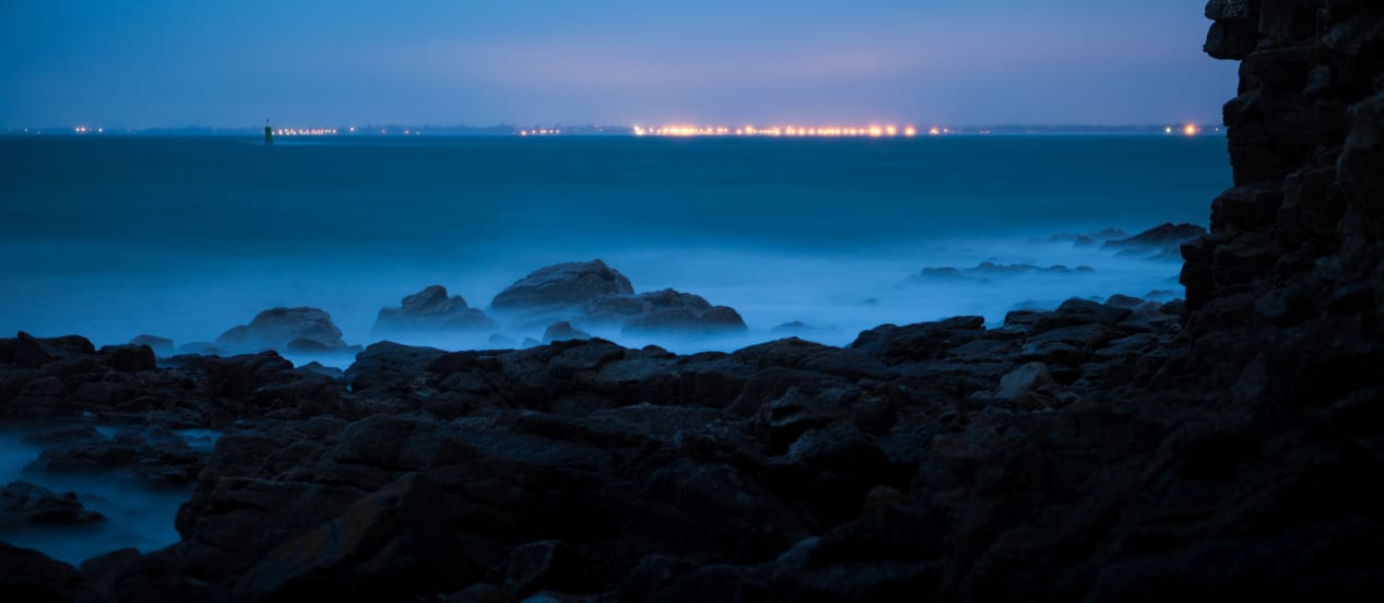 Nighttime seascape showing misty waters with rocky shore in the foreground and distant city lights across the water.