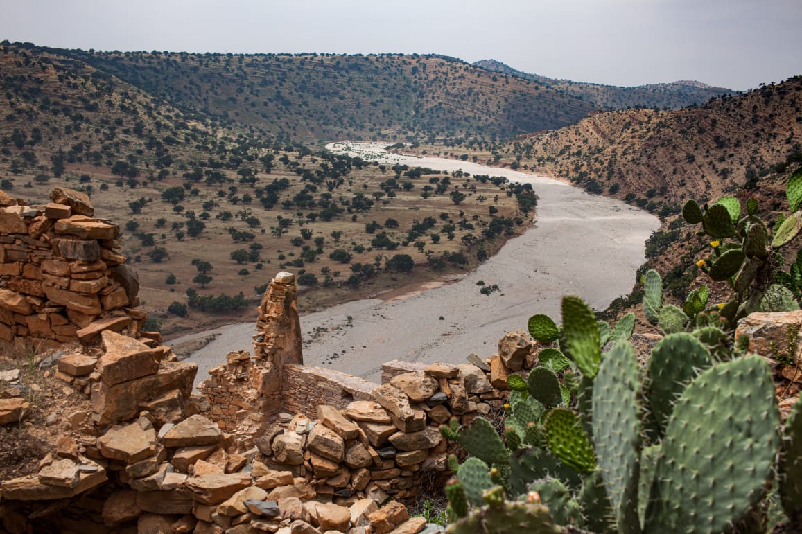 Rugged terrain with a dry river bed winding through hills, cactus in the foreground, and remnants of stone walls.