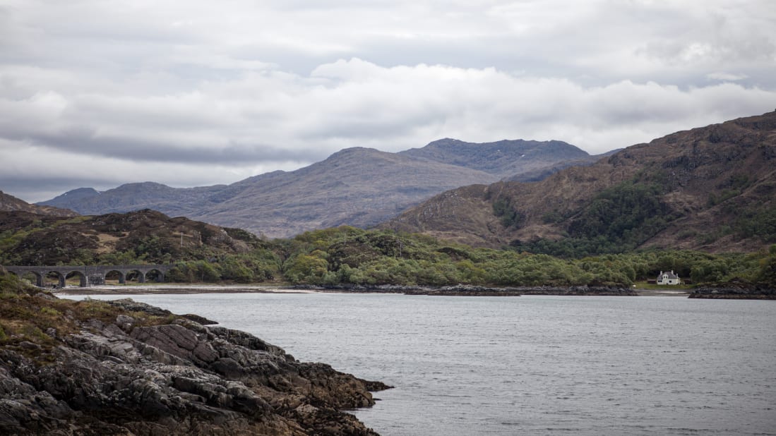 A coastal landscape showing rocky shores in the foreground, a calm sea, a viaduct overlooking water mid-scene, mountainous terrain, and cloudy skies. A small white building is nestled among trees near the water's edge.