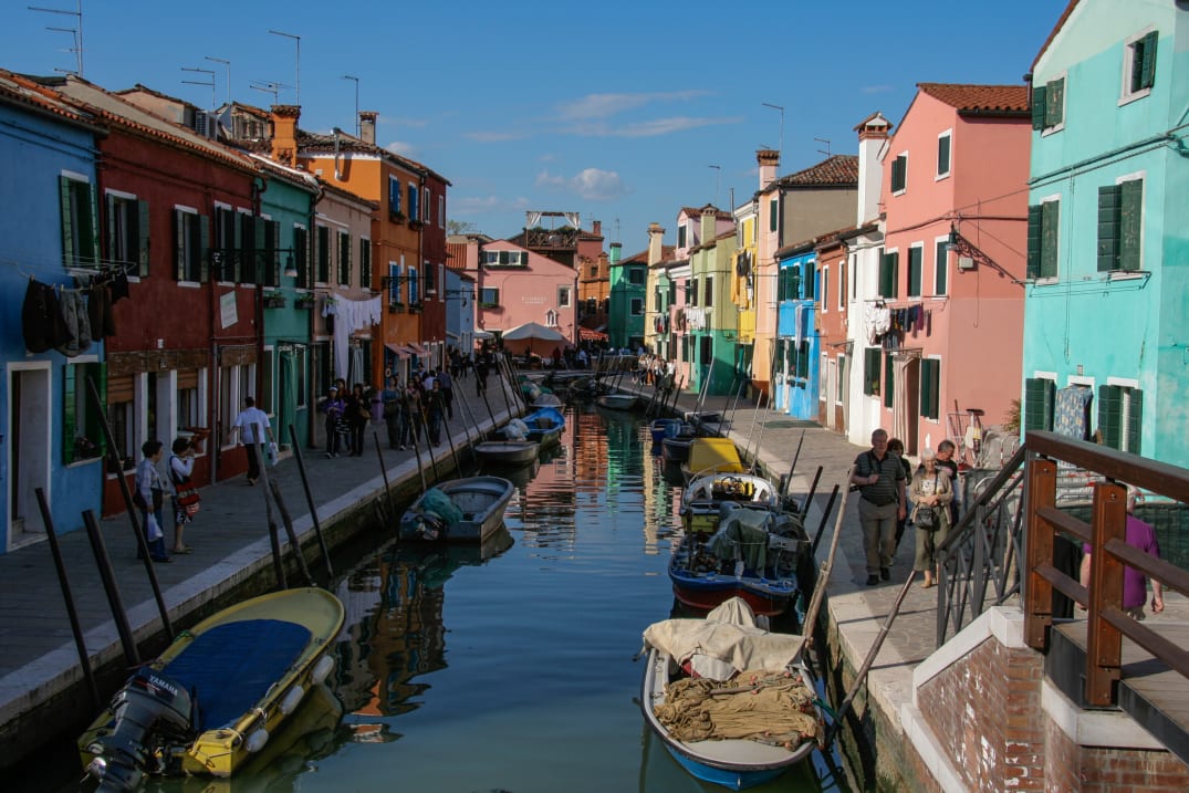 Colorful buildings line a narrow canal with boats in Burano, Italy, as people walk along the sidewalks.