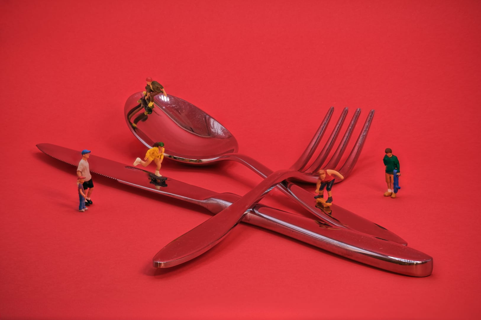 A creative scene featuring miniature figurines on a set of polished cutlery, placed against a red background. The figurines appear to use the cutlery as a skate park.