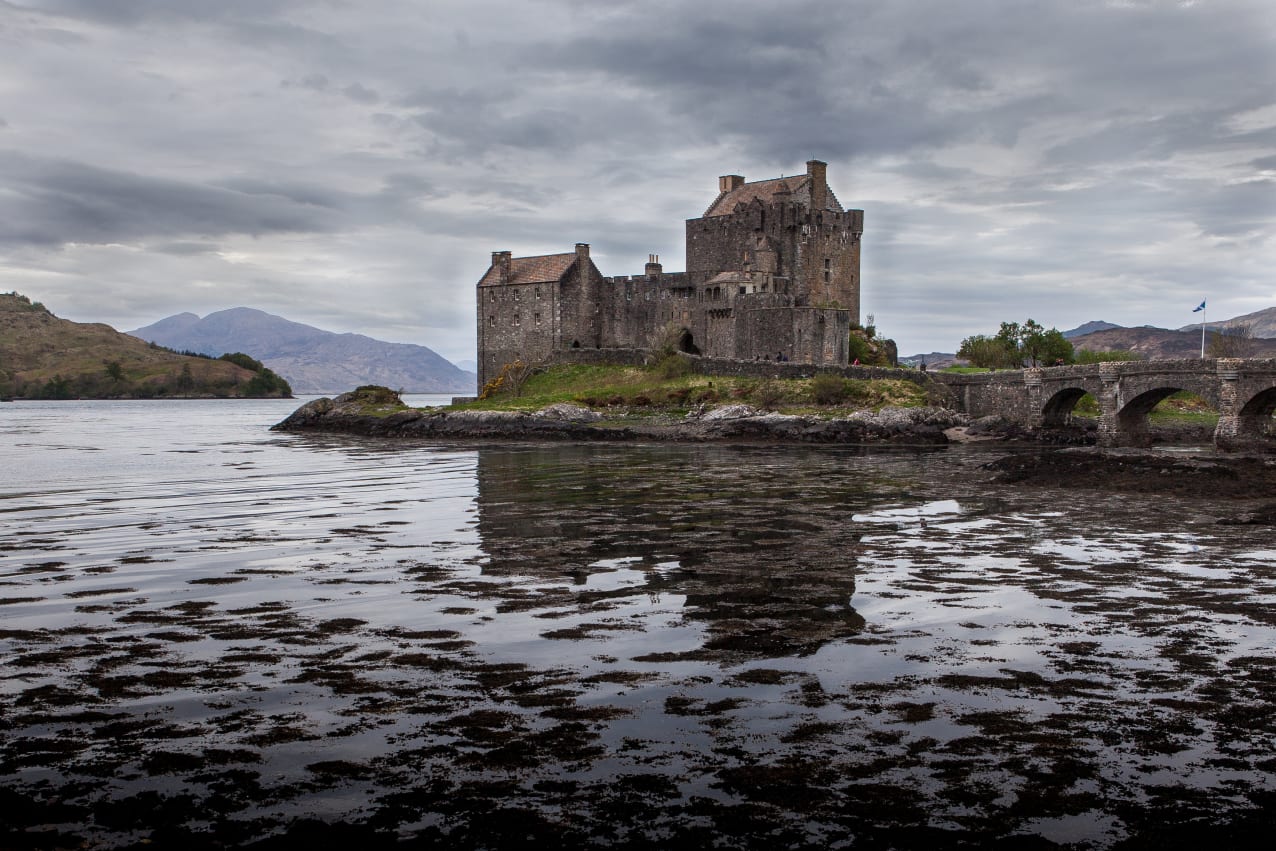 An ancient castle on a water's edge with a stone bridge, surrounded by mountains under a cloudy sky.