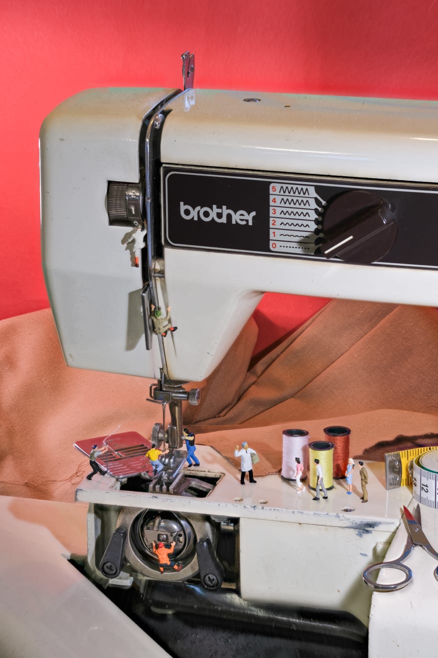 A vintage sewing machine is crowded with several miniature figures engaged in various activities around it, apparently workers working on it, and people interested in clothes. Spools of thread in various colors are laid out nearby, along with a tape measure and scissors.