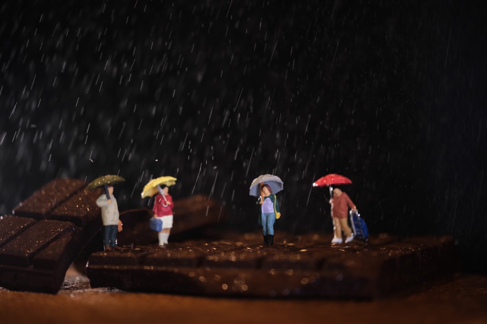 Four miniature figures holding umbrellas stand on a piece of chocolate while rain falls around them.
