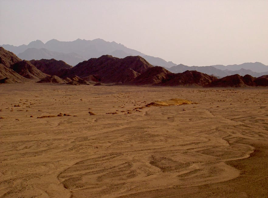 Desert landscape with sandy ground and rocky hills under a hazy sky.