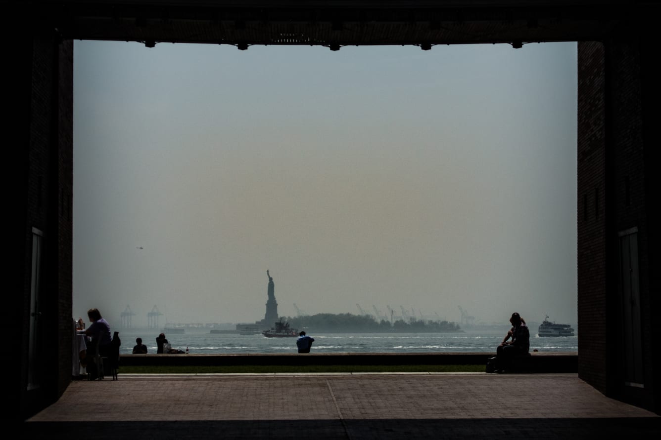 A framed view from a dark interior space looking out to the Statue of Liberty, with people sitting and walking in the foreground.