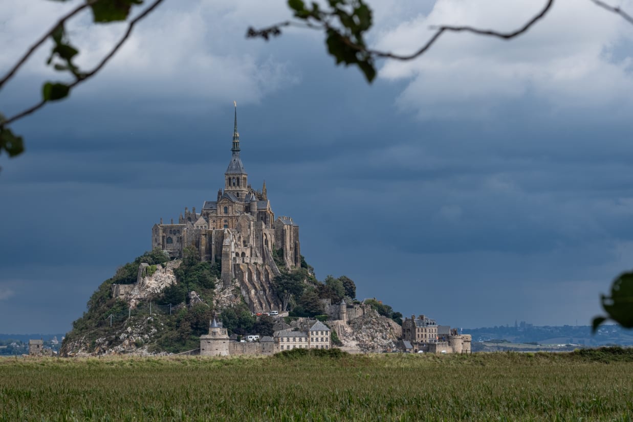 Mont Saint-Michel, a historic island commune with a monastery, against a dramatic cloudy sky, framed by foliage.
