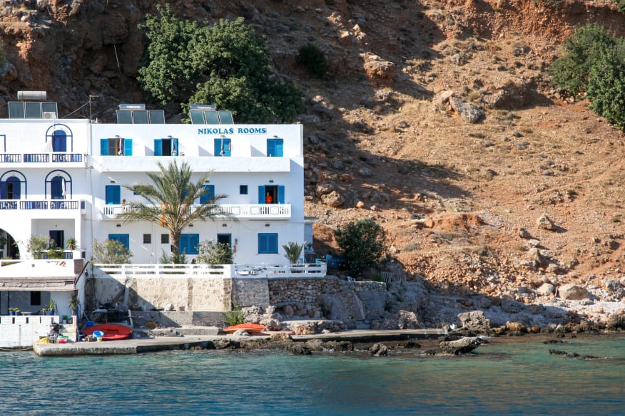 White building labeled "Nikolas Rooms" by the waterfront, with blue shutters and doors, against a rocky hillside and clear blue water in the foreground.