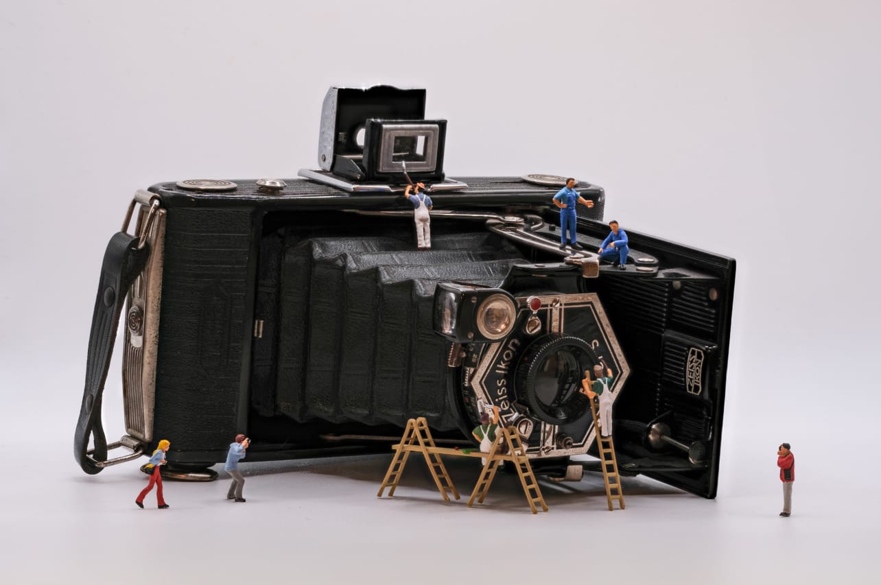 A large vintage camera is depicted, surrounded by miniature figures who are climbing ladders, performing maintenance, and taking photos. The scene combines elements of photography and creativity, showcasing the interaction between the miniatures and the camera.