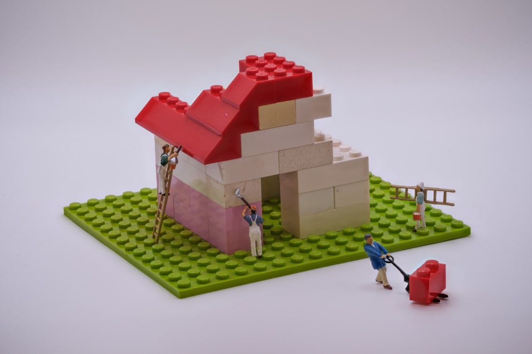 A colorful Lego house under construction, featuring a red roof and various bricks. Miniature figures are working: one on a ladder painting the house, another using a tool, and a third pushing a wheelbarrow.